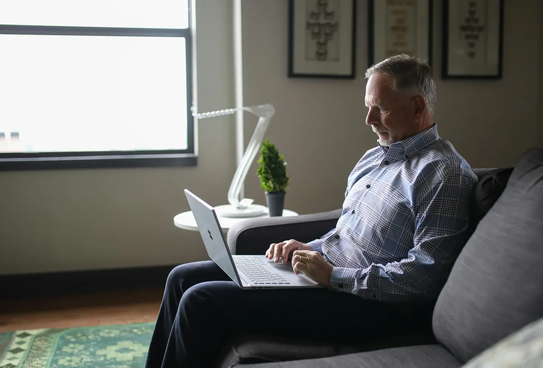 A man in his fifties on a sofa at home, laptop open for an appointment